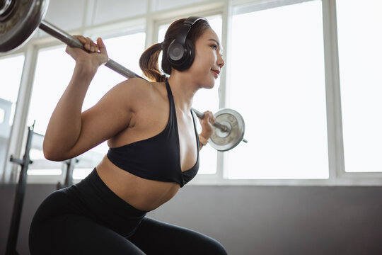 squat barbell training strength athlete Asian woman performing a barbell squat while wearing headphones in a modern gym highlighting strength training fitness motivation and a healthy lifestyle
