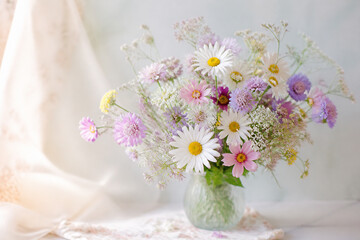 Obraz premium A bouquet of mixed wildflowers in a glass vase sits on a light surface near draped fabric.