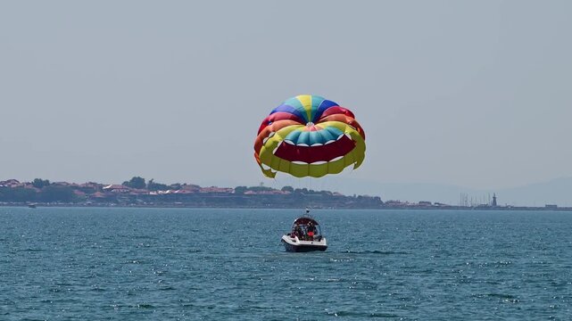 A vibrant, multi-colored parasail is towed closely behind a motorboat on a bright blue sea, with a distant coastal town featuring red-roofed buildings along the horizon.