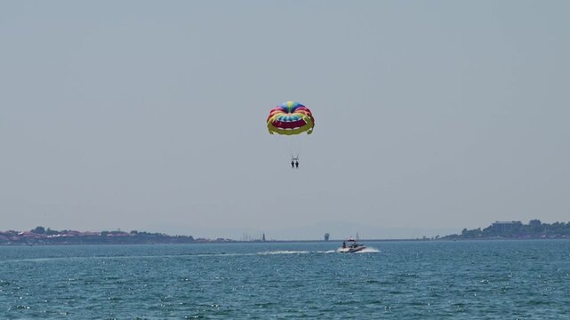 Two people enjoy a thrilling tandem parasailing ride under a brightly colored parachute, towed by a speedboat across a calm blue sea with a distant coastline in the background.