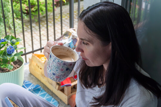 Young brunette woman enjoying a cup of coffee on a peaceful balcony decorated with blooming blue hydrangea (Hydrangea macrophylla). Cozy morning atmosphere, home relaxation, and comfortable everyday l