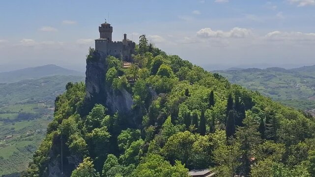 View of the historic Guaita Tower and the medieval city of San Marino perched atop Mount Titano, surrounded by lush green forests and clear blue sky in San Marino.
