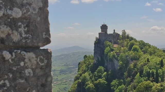 View of the historic Guaita Tower and the medieval city of San Marino perched atop Mount Titano, surrounded by lush green forests and clear blue sky in San Marino.