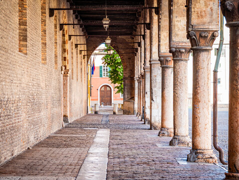 Historic arcade featuring a row of weathered stone columns and archways, lining a paved alley with ancient brick walls, leading to an old building and inviting exploration of architectural heritage