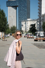 Fototapeta premium Woman looks back while walking along Shkolnaya Street in Moscow carrying light jacket on sunny summer day. Urban lifestyle, travel, city walk, confidence, leisure, warm weather, freedom