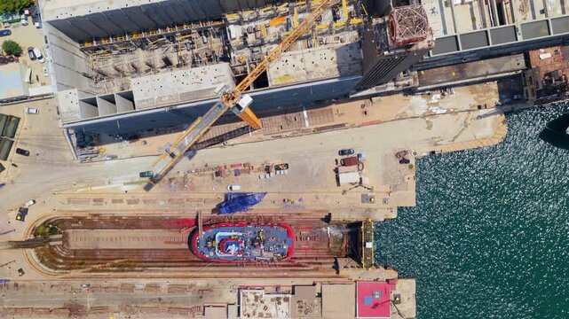 Top-down aerial perspective showing a large caisson gate slowly sliding out of a dry dock, uncovering a massive concrete ship hull under construction at an industrial shipyard by the sea