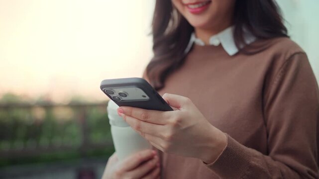 Close up of happy Asian businesswoman hands using smartphone while holding a tumbler outdoors at office balcony during sunset break, young female entrepreneur texting on mobile phone online app