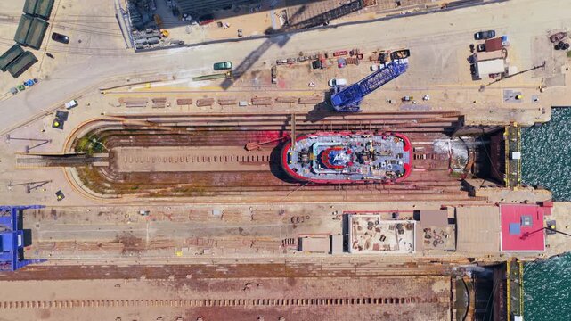 Industrial aerial view of a red tugboat launching from a shipyard's dry dock, sliding on rails into the sea for its maiden voyage after construction and maintenance in the harbor