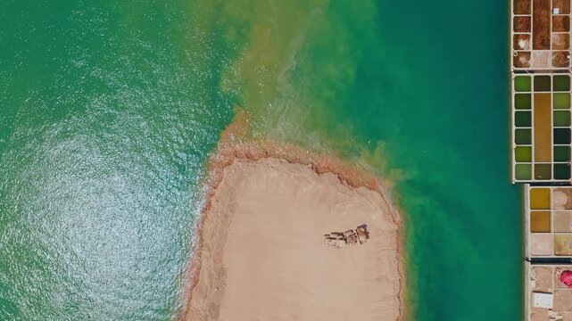 Aerial top-down view of a caravan of camels resting on a sandy spit of land in the dead sea, with vibrant evaporation ponds visible on the shore, showcasing a unique and tranquil scenery