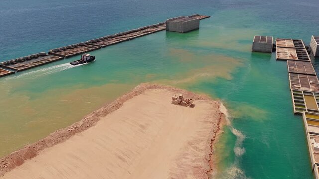Aerial view of a heavy bulldozer pushing sand into the turquoise water for a coastal land reclamation project, as a tugboat sails past with barges, creating a new man-made peninsula