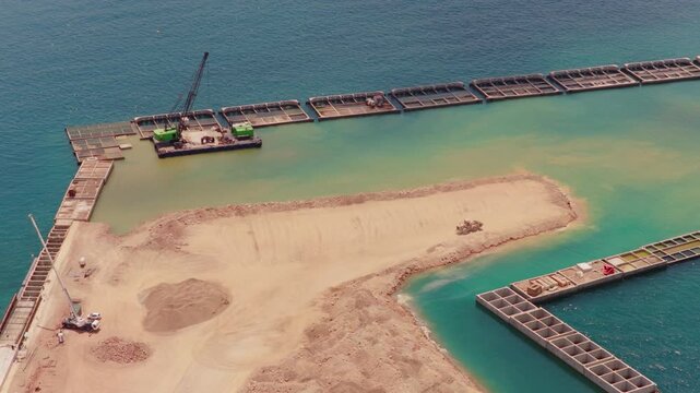 Heavy machinery working on a large-scale coastal engineering project, creating a new peninsula through land reclamation with sand and concrete caissons for a future marina or port