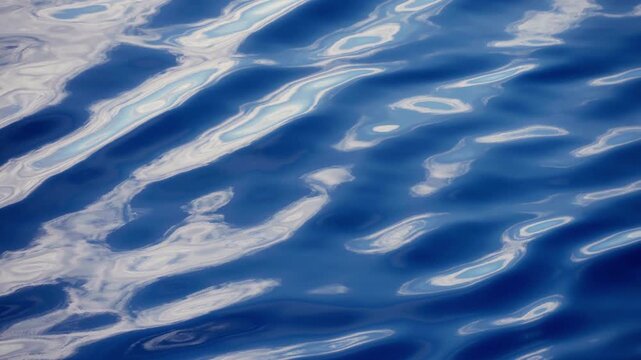 Serene blue ocean water surface featuring gentle ripples and soft waves creating abstract patterns. White sky reflections dance across the tranquil deep blue sea in calming motion.