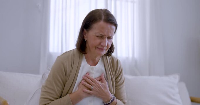 Elderly woman sits on living room sofa, pressing chest, leaning forward and lifting head while wincing from pain, expressing distress, health concern, and need for attentive support at home.