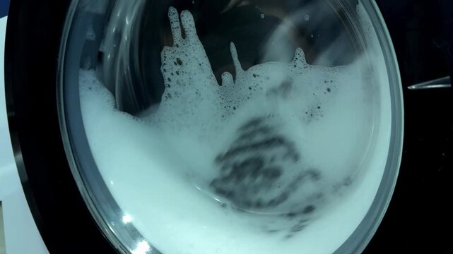 Video of clothes spinning at high speed inside an automatic washing machine drum with soapy water waves.