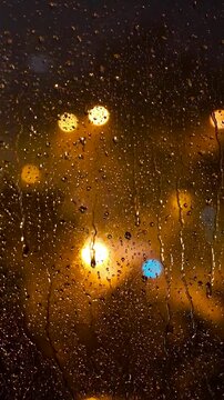 Vertical Raindrops on Glass Window with Warm Night City Bokeh and Thunderstorm Lightning. Detailed water drops on foreground with warm orange, yellow, and red blurry bokeh lights in the background.