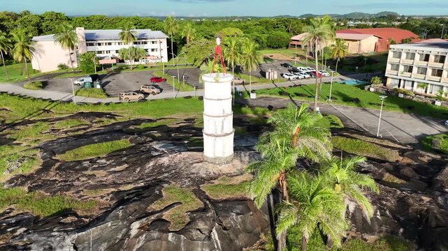 Smooth morning drone pull-away from the historic Tour Dreyfus, gradually revealing the rocky coastline, palm trees, nearby buildings and the wider landscape of Kourou, French Guiana, France.
