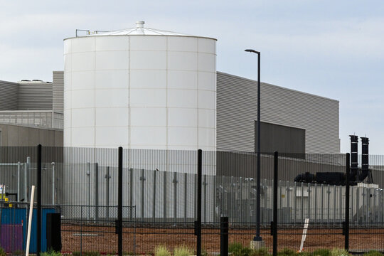 Large water storage tank at a hyperscale data center viewed through a security perimeter fence, representing cooling infrastructure, secure digital facilities, and cloud computing operations