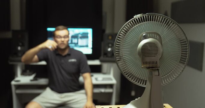 A man sits in front of an electric fan, shielding his face from the heat while trying to cool down in a warm home office.