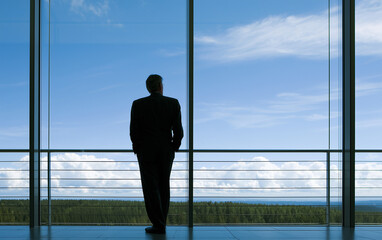 Fototapeta premium Businessman in suit looking out expansive office window at bright blue sky with clouds