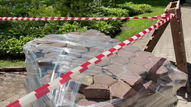 Red and white warning tape blocking a pallet of paving stones wrapped in plastic film outdoors. Road repair