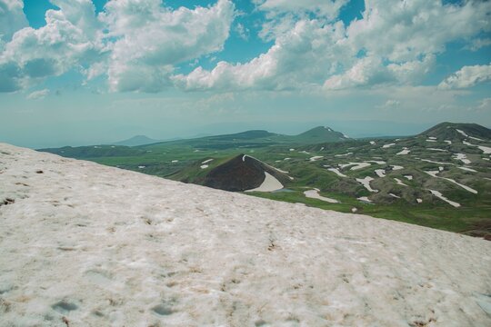 Panoramic Landscape of Volcanic Plateau with Red Earth Cone, Patchy Snow, and Fluffy Cumulus Clouds
