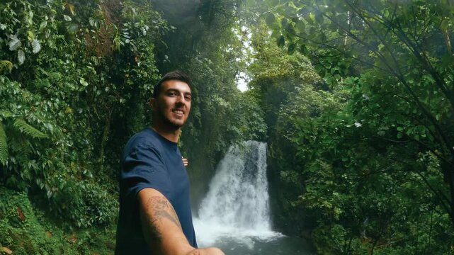 Male tourist filming himself with a selfie stick at a waterfall in the rainforest. Young man enjoying nature in Arenal Volcano National Park