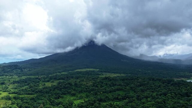 Majestic Arenal Volcano in La Fortuna, Costa Rica, with clouds swirling around its peak and casting dynamic shadows over the lush rainforest below