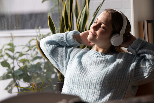 Young woman relaxing with eyes closed, wearing white headphones and cozy sweater, stretching arms behind head near indoor plants at home