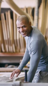 Confident craftsman working in his wood shop. Bald artisan sanding a wooden surface by hand, looking at the camera and smiling proudly