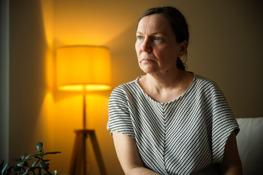 Middle-aged woman deep in thought while sitting in a cozy living room. Reflection, problem solving, concern and emotional wellbeing theme.