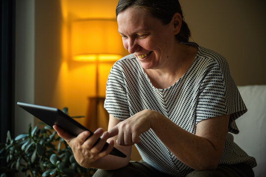 Smiling woman using a tablet computer in a comfortable home setting during the evening. Digital lifestyle, remote work and online communication concept.