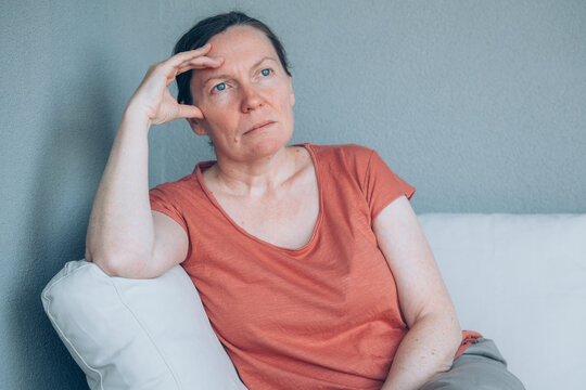 Middle-aged woman sits on a sofa, gazing thoughtfully and appearing pensive and reflective. Calm indoor scene conveying contemplation.