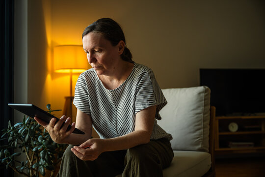 Middle-aged woman using a digital tablet while relaxing at home in the evening. Online communication, remote work and modern technology lifestyle concept.