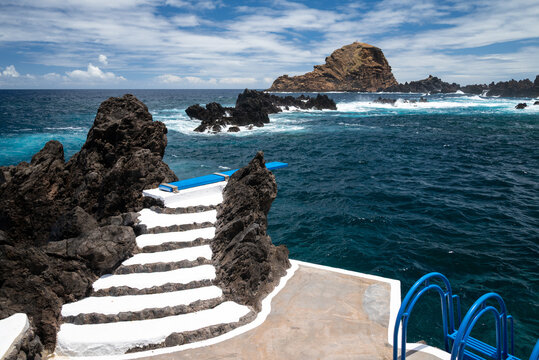 White-painted steps and a blue diving board leading into the natural volcanic swimming pools of Porto Moniz, with the Ilheu Mole islet beyond, north-west Madeira, Portugal.