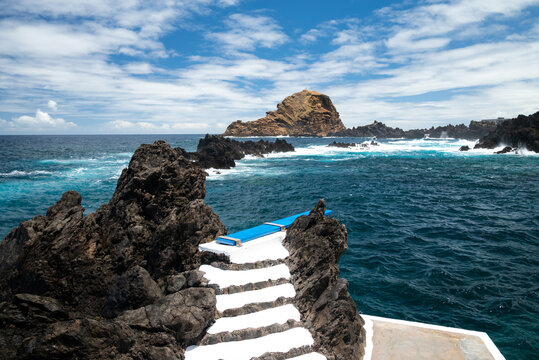 Whitewashed steps descend past rugged lava rock to the natural sea pools of Porto Moniz, with the Ilheu Mole islet on the horizon, north-west coast of Madeira, Portugal.