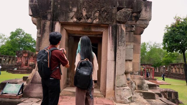 Young Asian backpackers walking and exploring historical stone temple site on sunny day. Travel destination adventure, historic archaeology discovery and summer vacation trip.