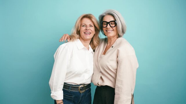 Two cheerful elderly women with beautiful smiles embracing and looking at the camera. Best friends sharing a moment against a studio background