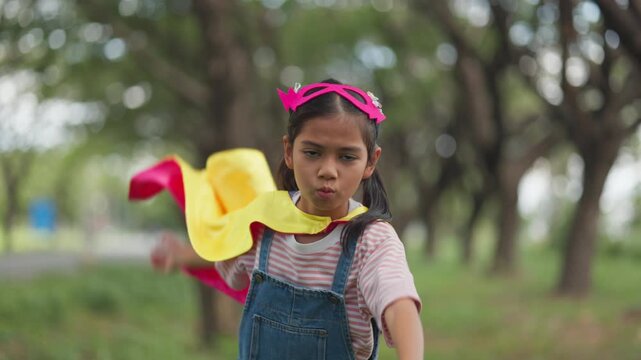 Cute Asian children playing superhero roleplay with colorful cape and mask running in outdoor green park. Kids imagination, creative future dream, and brave hero inspiration.