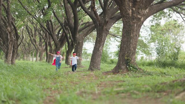 Active Asian children wearing superhero cape and mask having fun running in tree tunnel park. Kids imagination, creative future dream, and brave hero inspiration activity.
