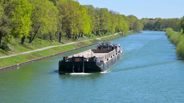 A bulk cargo barge ship navigates along a scenic inland water canal lined with green trees in Germany. 4K slow motion footage