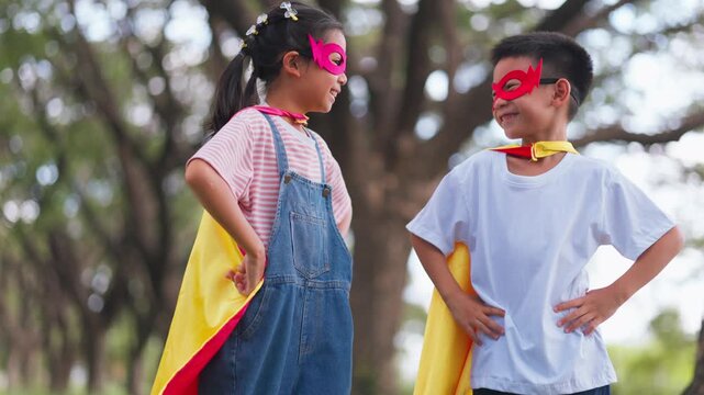 Cute Asian children playing superhero roleplay with colorful cape and mask running in outdoor green park. Kids imagination, creative future dream, and brave hero inspiration.