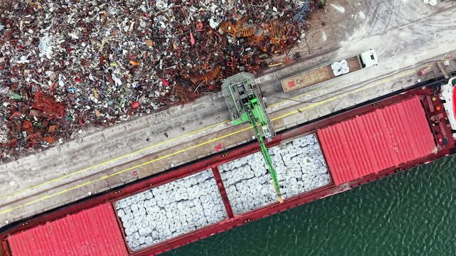 A cargo ship is being loaded with materials from a scrapyard located next to the water. Workers and equipment are moving items onto the ship while it is docked.