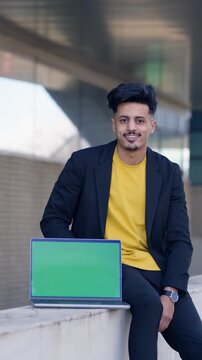 Young moroccan businessman wearing a black jacket and a yellow t shirt sitting outdoors showing a laptop with a green screen. Vertical