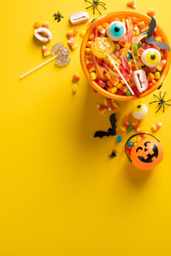 Happy Halloween candy and pumpkin bucket with lollipops gummy worms and spooky decorations on vertical vivid yellow background