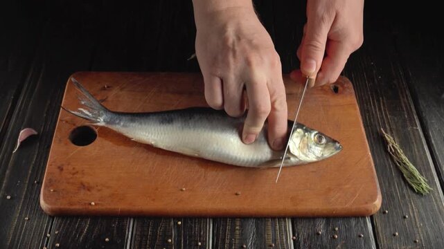 Hands of a person skillfully filleting a fish on a wooden cutting board, showcasing the precise cutting technique and the herring fish's body positioned for easy access during the preparation process