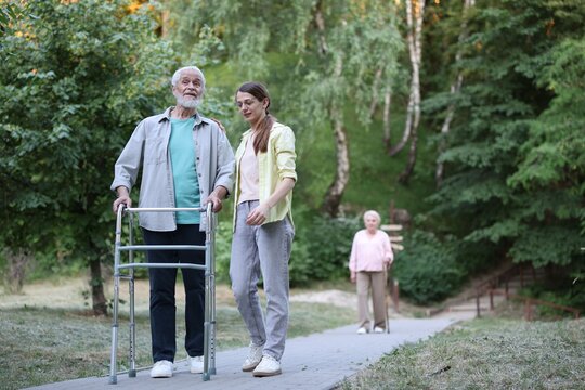 Caregiver supporting elderly man with walking frame and senior woman outdoors, selective focus