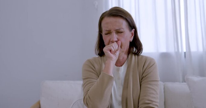 Senior woman sits on living room sofa, holding chest and repeatedly coughing into hand, expressing sudden throat discomfort, health concern, respiratory symptom, and need for caring support at home.