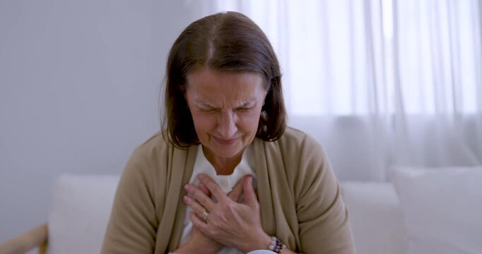 Senior woman sits on living room sofa, clutching chest with both hands, bending forward and leaning back while grimacing from sudden pain, expressing health concern and need for attentive home care.