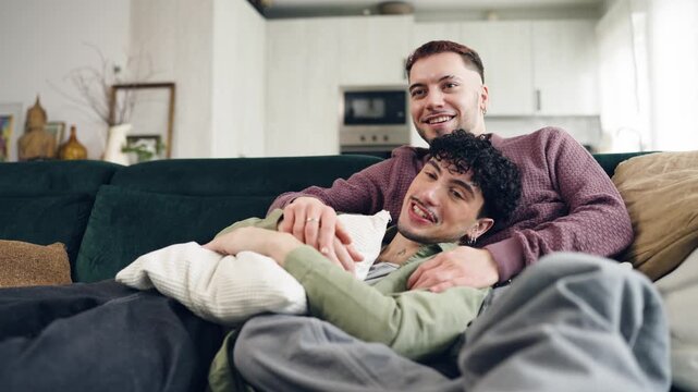 Loving gay couple relaxing together on the sofa at home, watching a movie. Intimate and cozy moment of two men cuddling in their living room