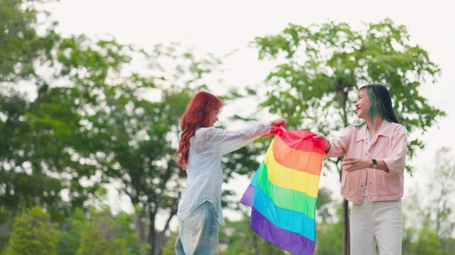 Asian LGBTQ women couple sharing happy moments and holding pride flag outdoors in park. Inclusive community, equality movement, diversity, and joyful relationship lifestyle.